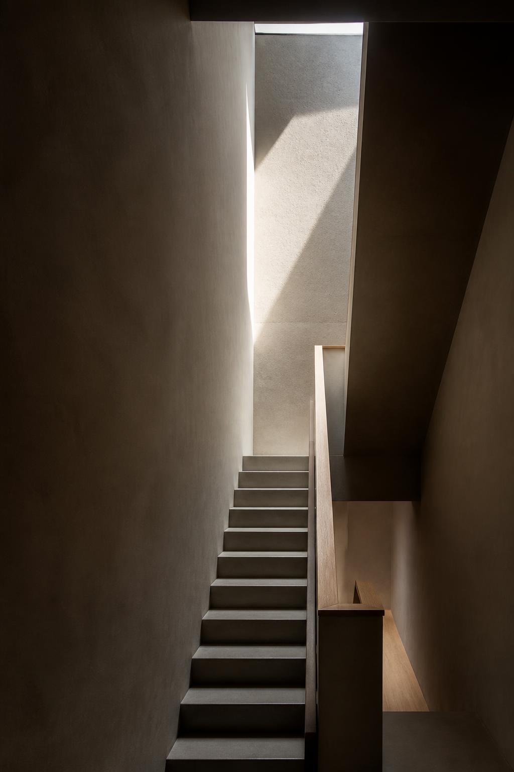 Concrete stair void lit by a narrow shaft of daylight with an oak handrail